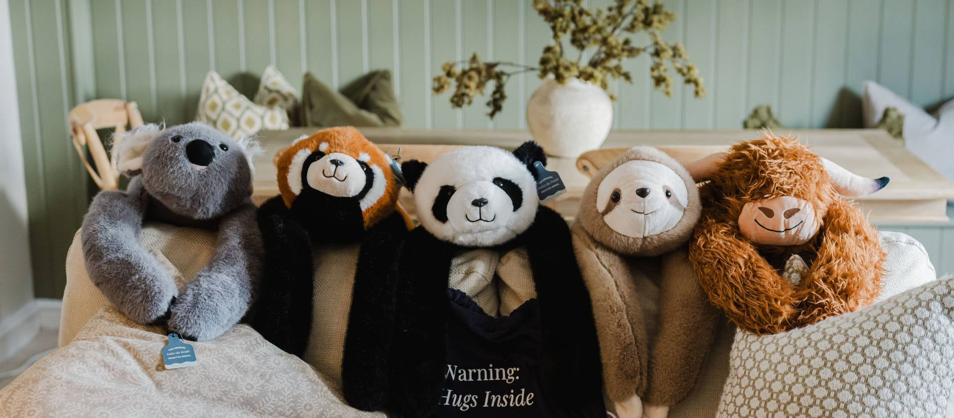 Five plush animal toys arranged on a beige sofa in a styled interior setting. The toys include a gray bear, a red panda, a panda in a black outfit with visible warning text reading 'Warning: Augs Inside' and 'PULSE OF POTENTIAL', a tan sloth, and a brown alpaca.
